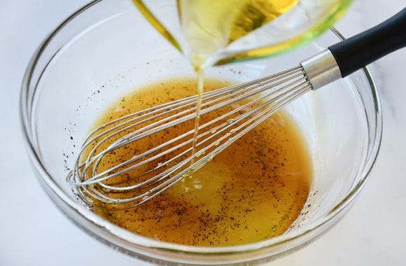 Olive oil being poured into a clear bowl containing dressing and a whisk