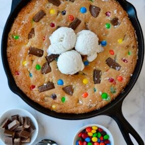 Leftover Halloween Candy Skillet Cookie topped with vanilla ice cream next to small bowls containing chocolate candy