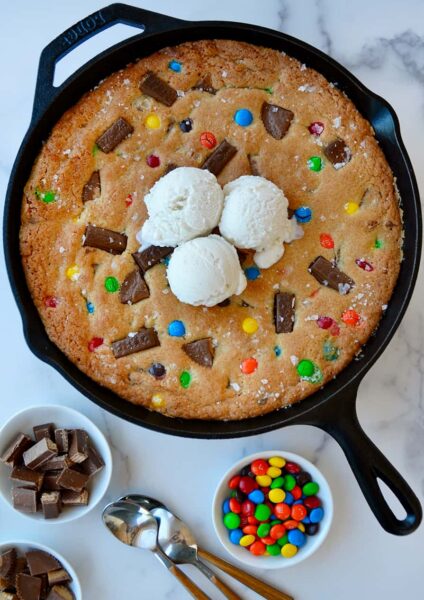 Leftover Halloween Candy Skillet Cookie topped with vanilla ice cream next to small bowls containing chocolate candy