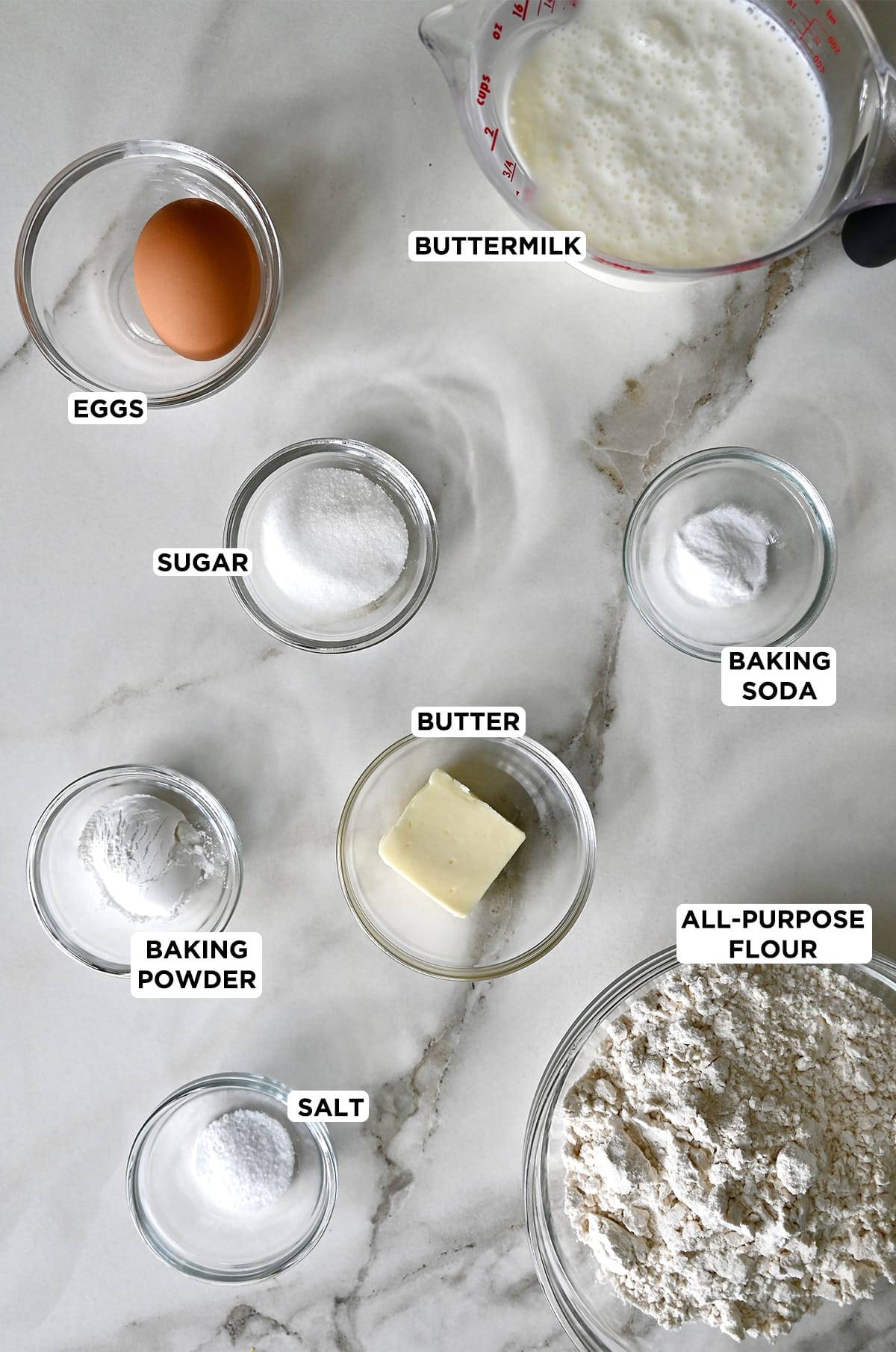 Various sizes of glass bowls containing these ingredients: buttermilk, baking soda, all-purpose flour, butter, salt, baking powder, sugar and a whole egg.