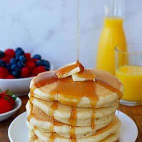 A tall stack of buttermilk pancakes dripping with maple syrup and topped with two pads of butter. Orange juice and fruit are in the background.