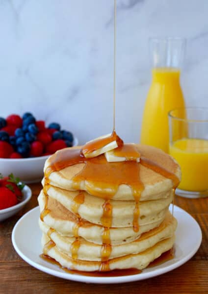 A tall stack of buttermilk pancakes dripping with maple syrup and topped with two pads of butter. Orange juice and fruit are in the background.