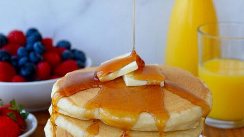 A tall stack of buttermilk pancakes dripping with maple syrup and topped with two pads of butter. Orange juice and fruit are in the background.