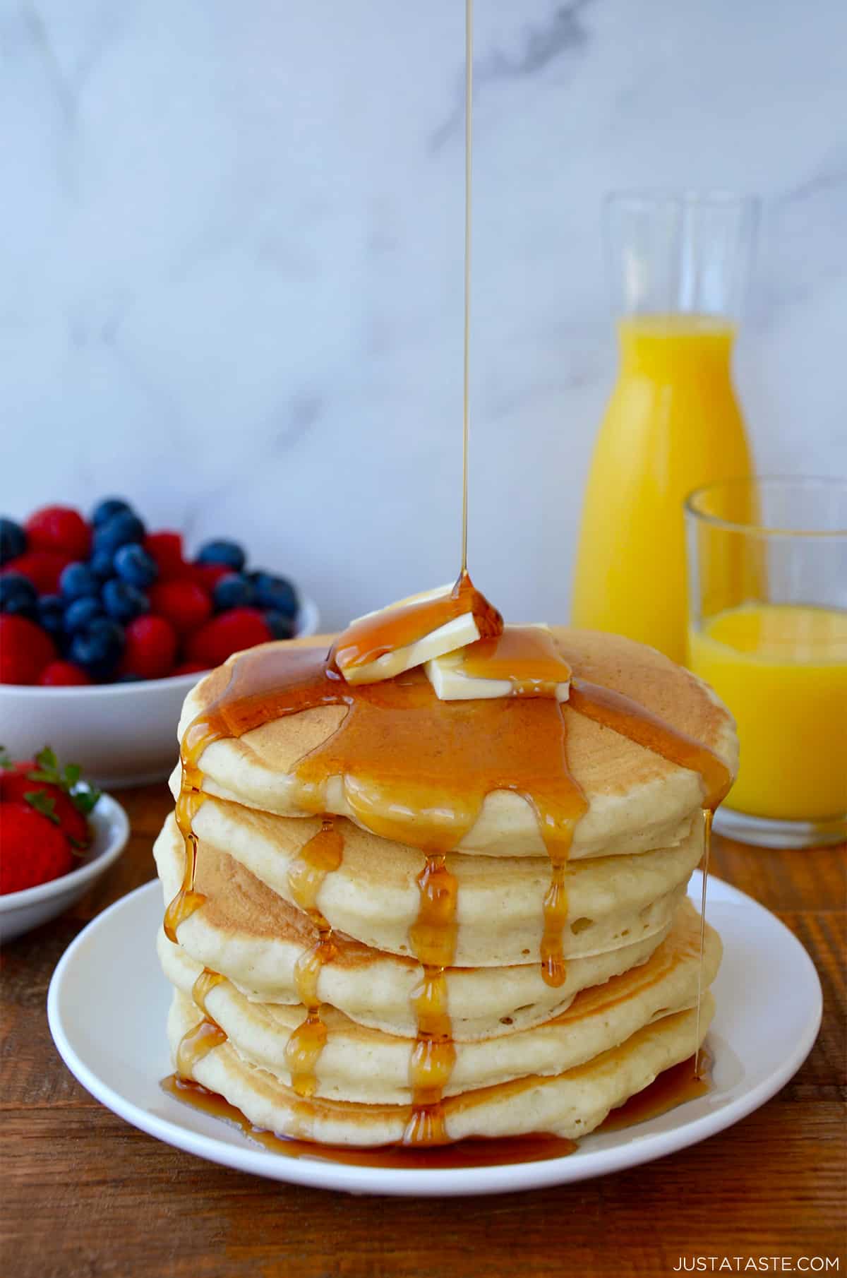 A tall stack of buttermilk pancakes dripping with maple syrup and topped with two pads of butter. Orange juice and fruit are in the background.