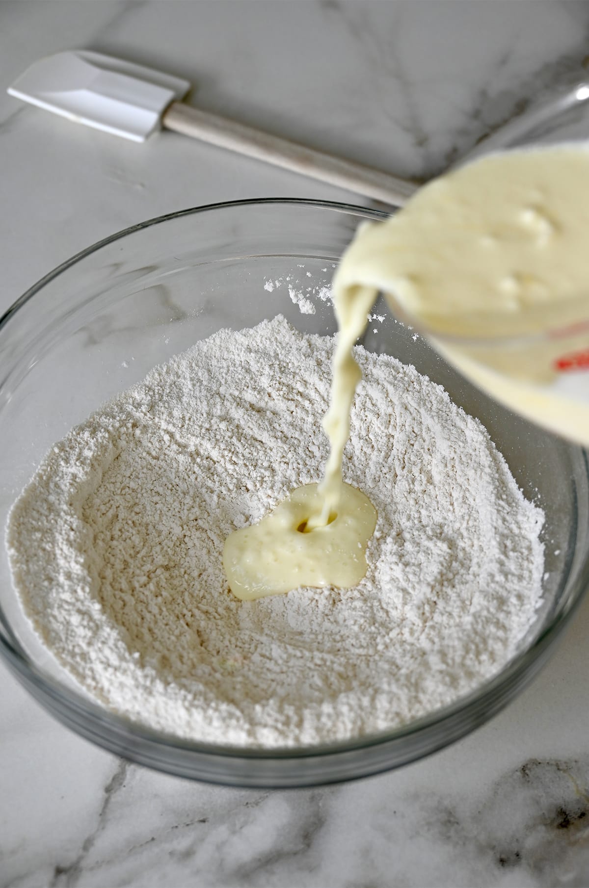 Buttermilk from a liquid measuring cup being poured atop flour in a glass bowl.