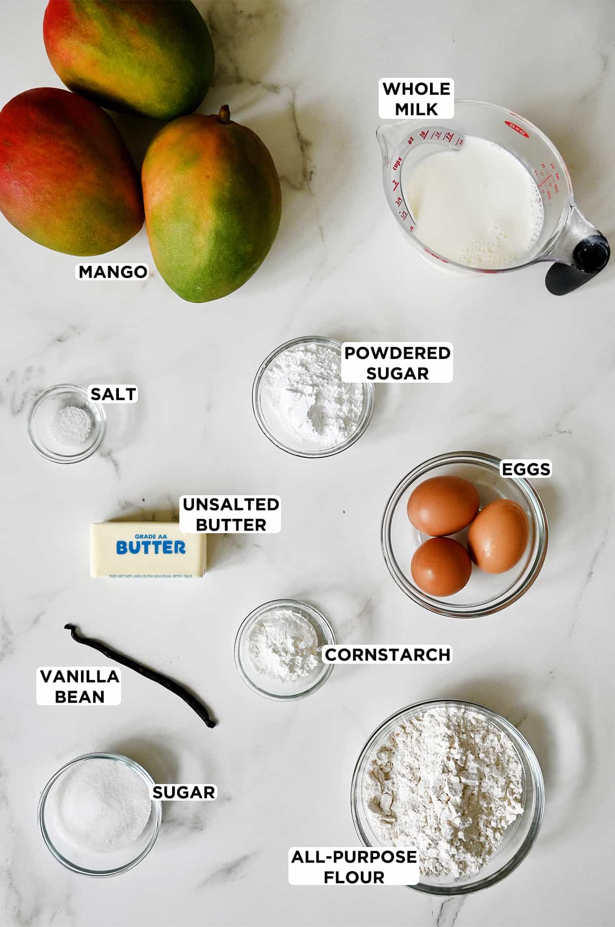 Three ripe mangoes next to a liquid measuring cup filled with whole milk and various sizes of glass bowls containing powdered sugar, eggs, a stick of unsalted butter, salt, a vanilla bean, flour and cornstarch.