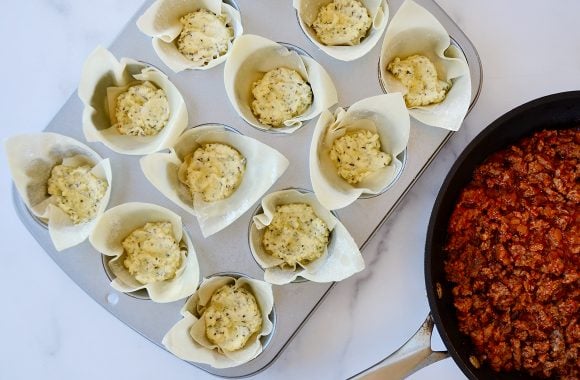 Muffin pan with wonton wrappers filled with ricotta cheese mixture next to skillet with Italian sausage