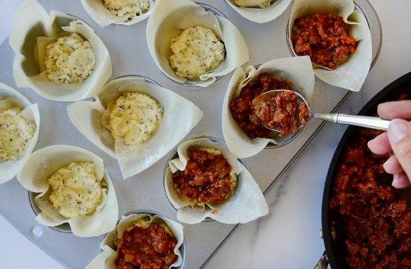 Hand holding spoon with sausage filling over pan with wonton wrappers filled with cheese