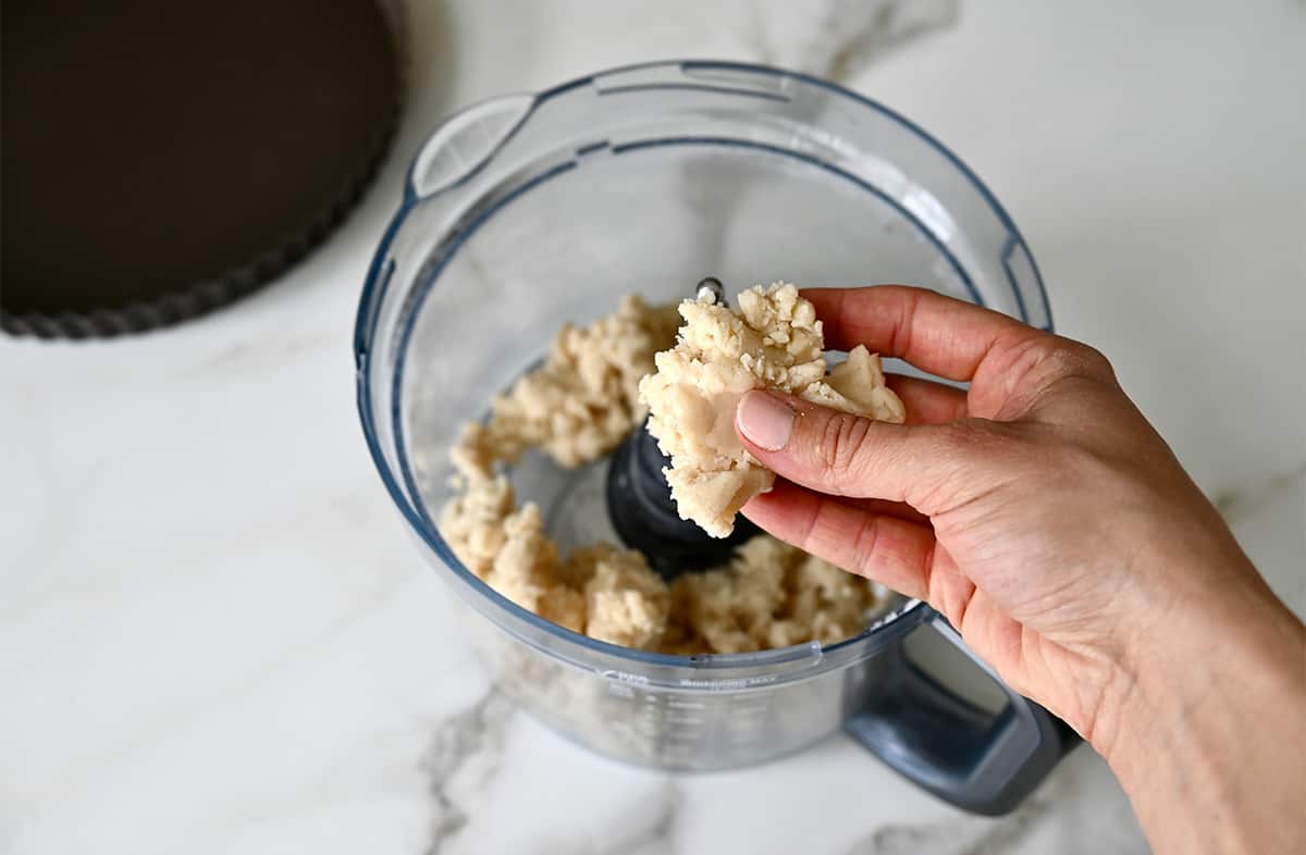 A hand holds shortbread dough above a food processor.
