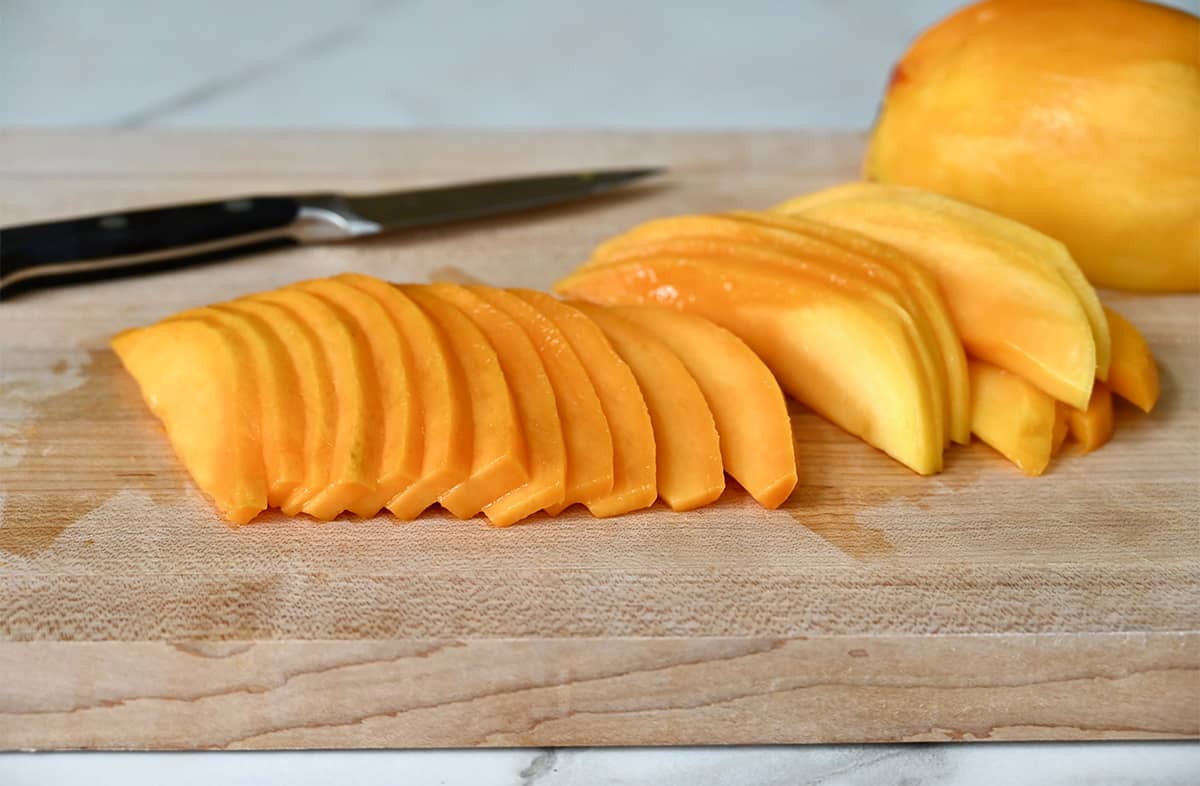 Thin slices of mango on a cutting board next to a sharp knife.