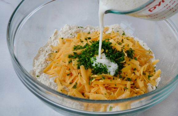 Buttermilk being poured into clear bowl containing flour, cheddar cheese and chives 