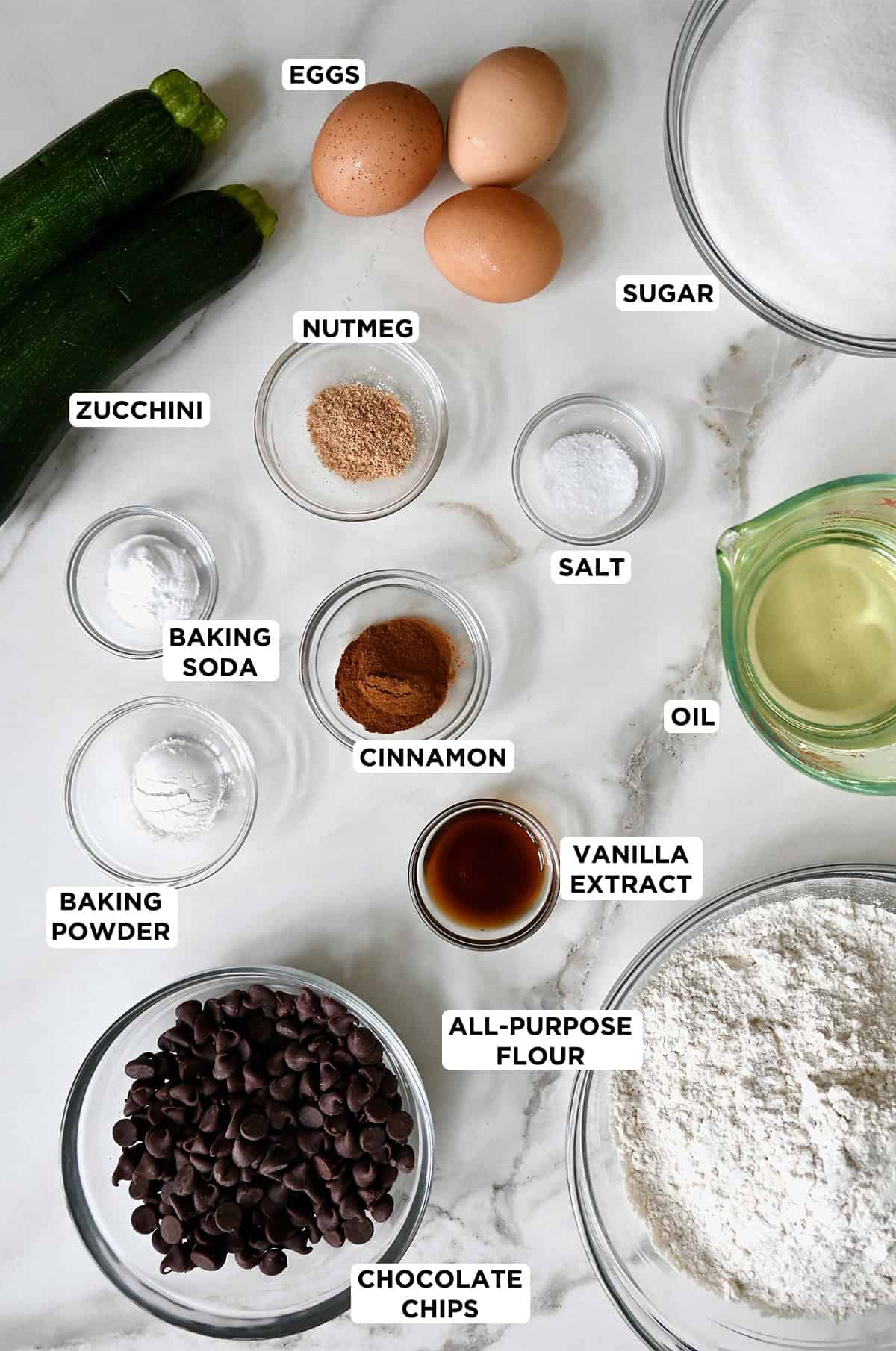 Ingredients for zucchini bread with chocolate chips in glass bowls, including eggs, sugar, nutmeg, cinnamon, salt, baking soda, vanilla extract, baking powder, flour, chocolate chips and two zucchini.