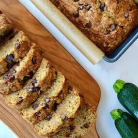Two loaves of moist chocolate chip zucchini bread, one in a loaf pan and one cut into perfect slices on a cutting board.