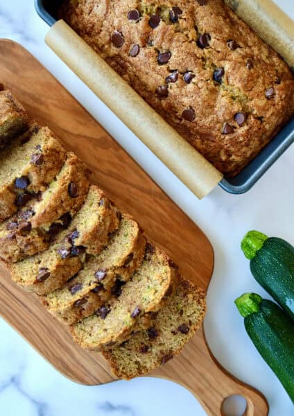 Two loaves of moist chocolate chip zucchini bread, one in a loaf pan and one cut into perfect slices on a cutting board.