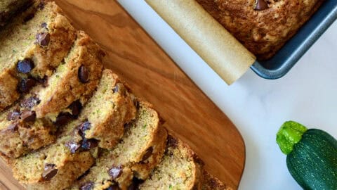 Two loaves of moist chocolate chip zucchini bread, one in a loaf pan and one cut into perfect slices on a cutting board.