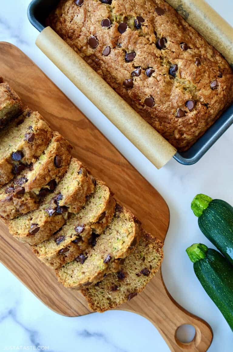 Two loaves of moist chocolate chip zucchini bread, one in a loaf pan and one cut into perfect slices on a cutting board.