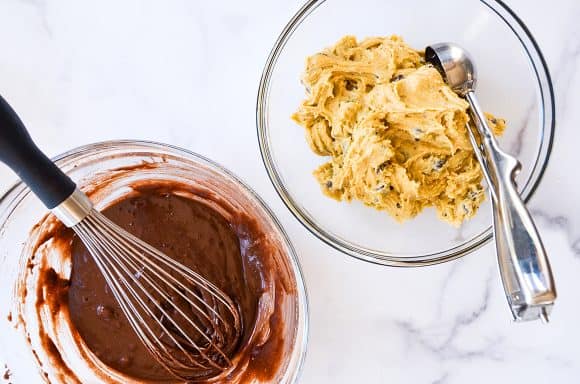 Two bowls, one containing cookie batter and cookie scoop, another containing brownie batter and whisk 