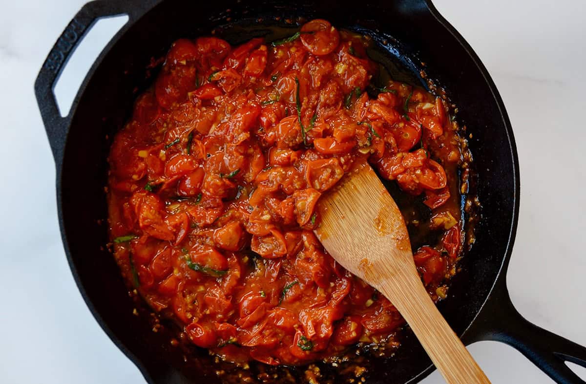 Jammy cherry tomato pasta sauce with fresh basil in a skillet with a wooden spoon.