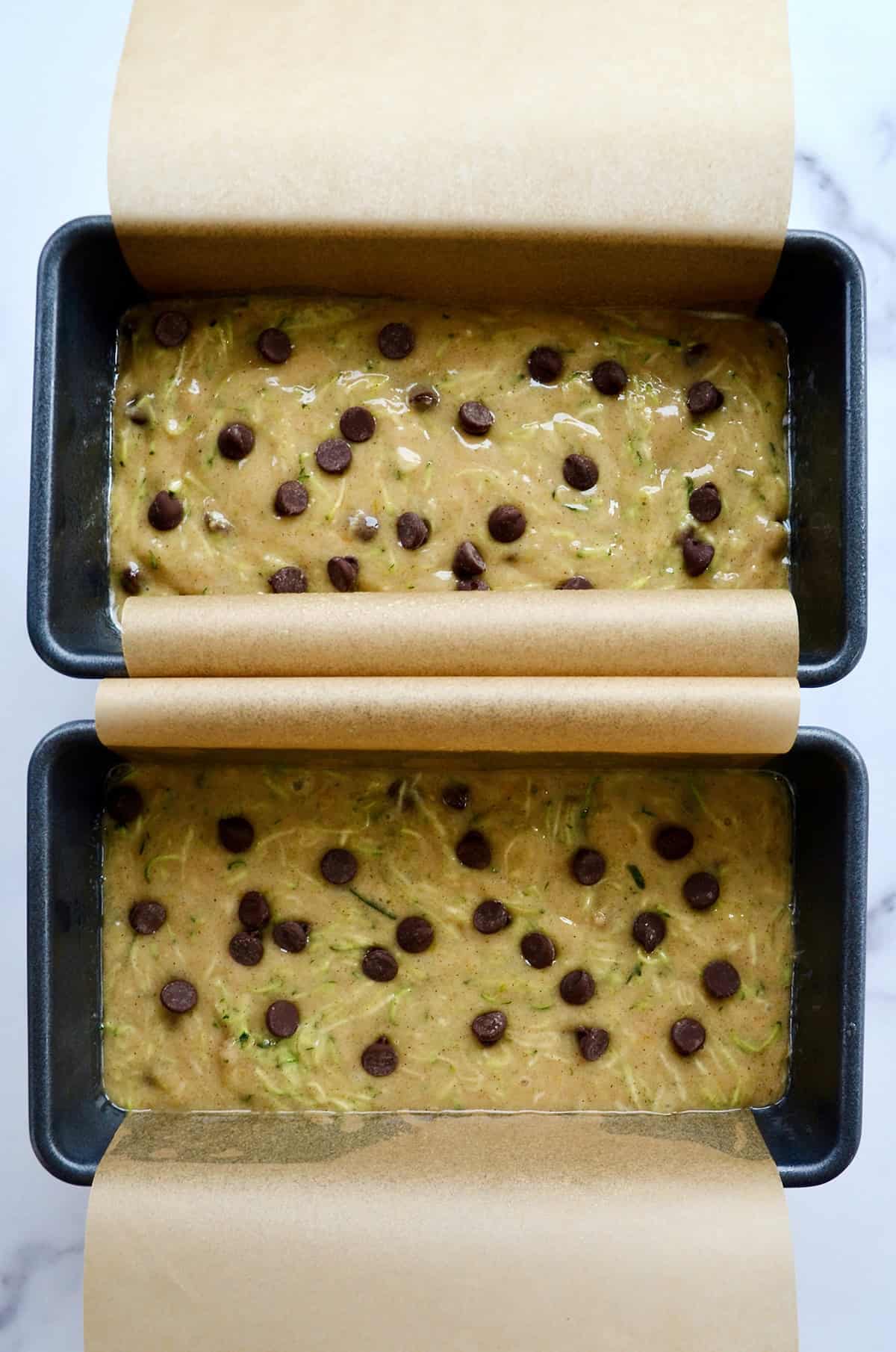 Two loaf pans lined with parchment paper filled with zucchini bread batter topped with chocolate chips.