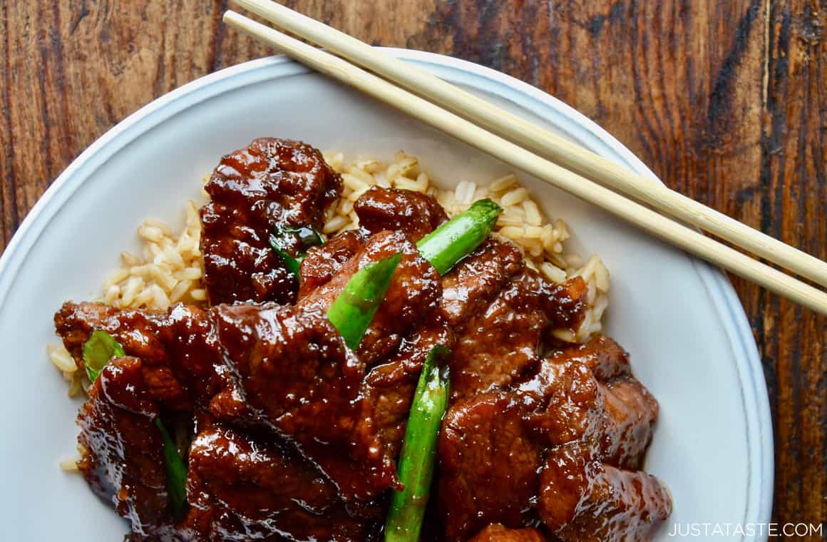 Top-down view of Mongolian beef garnished with slices of scallions over a bed of brown rice in a white serving bowl with chopsticks