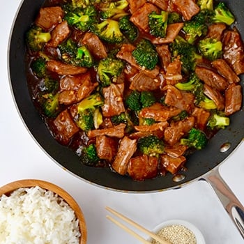 A skillet containing beef and broccoli with a bowl of white rice next to it