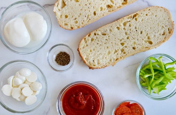 Halved bread next to various sizes of bowls containing green peppers, pepperoni, marinara sauce, mozzarella balls, Italian seasoning and burrata