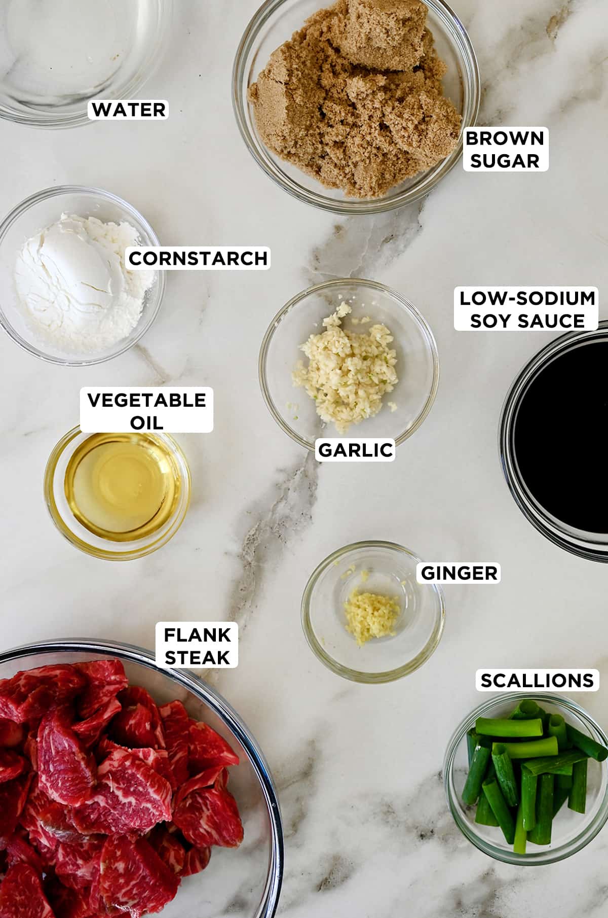 Various sizes of clear bowls containing ingredients to make Mongolian beef, including brown sugar, low-sodium soy sauce, scallions, minced ginger, minced garlic, thinly sliced flank steak, vegetable oil and cornstarch.