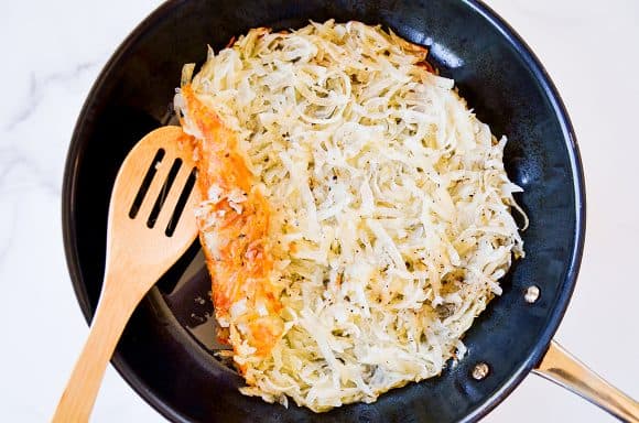 Large skillet containing crispy hash browns and wooden spoon
