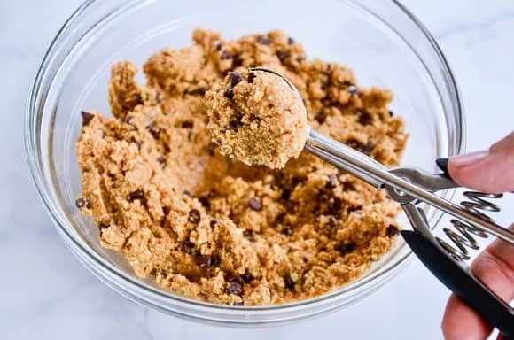 Hand holding small ice cream scoop over no-bake mixture in a clear bowl