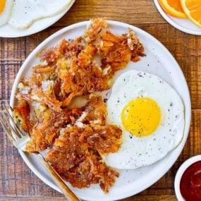 Top down view of a white plate containing the best crispy hash browns, a fork and a fried egg