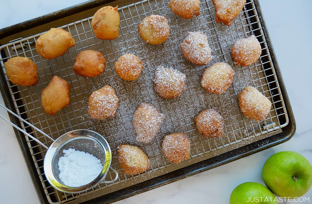 Apple fritters dusted with powdered sugar on a baking sheet with a wire cooling rack.