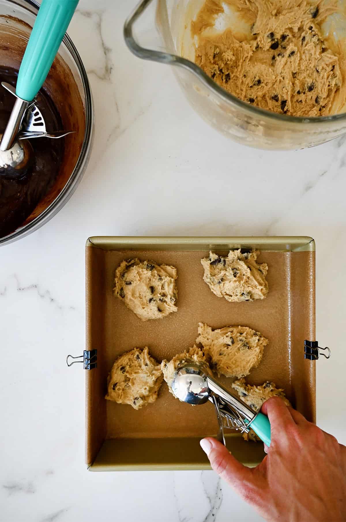 Using an ice cream scoop to add dollops of chocolate chip cookie dough to a lined square pan.