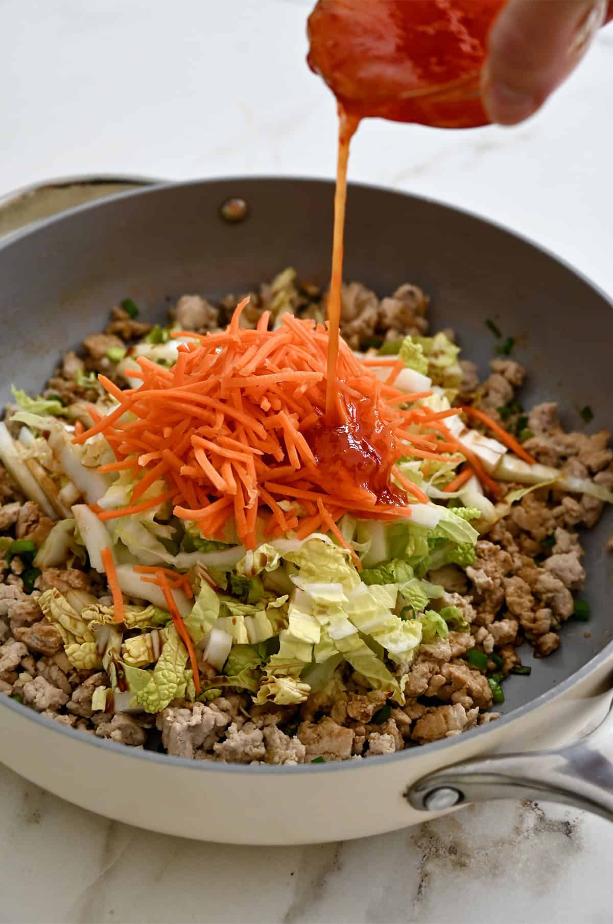 Shredded carrots, shredded cabbage and ground meat in a skillet being topped with sweet chili sauce.