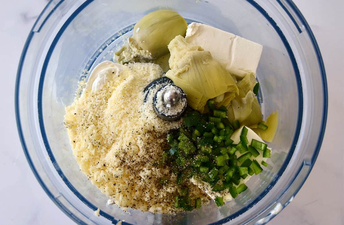 The bowl of a food processor containing grated Parmesan cheese, diced jalapeño, artichoke hearts, cream cheese, sour cream and mayo.