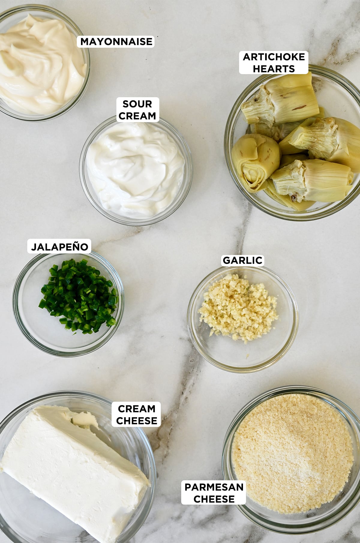 Various sizes of glass bowls containing ingredients for hot artichoke dip, including mayonnaise, sour cream, artichoke hearts, minced garlic, grated Parmesan cheese, cream cheese and diced jalapeños. 