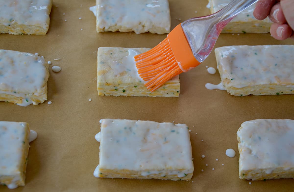 Rectangular unbaked biscuits on a sheet of parchment are brushed with heavy cream using a silicone brush