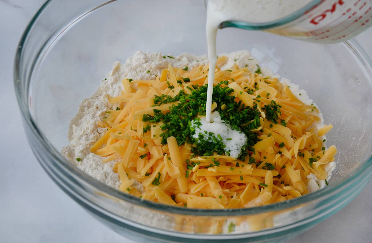 Buttermillk is being poured into a glass bowl containing flour, butter, cheese and chives.