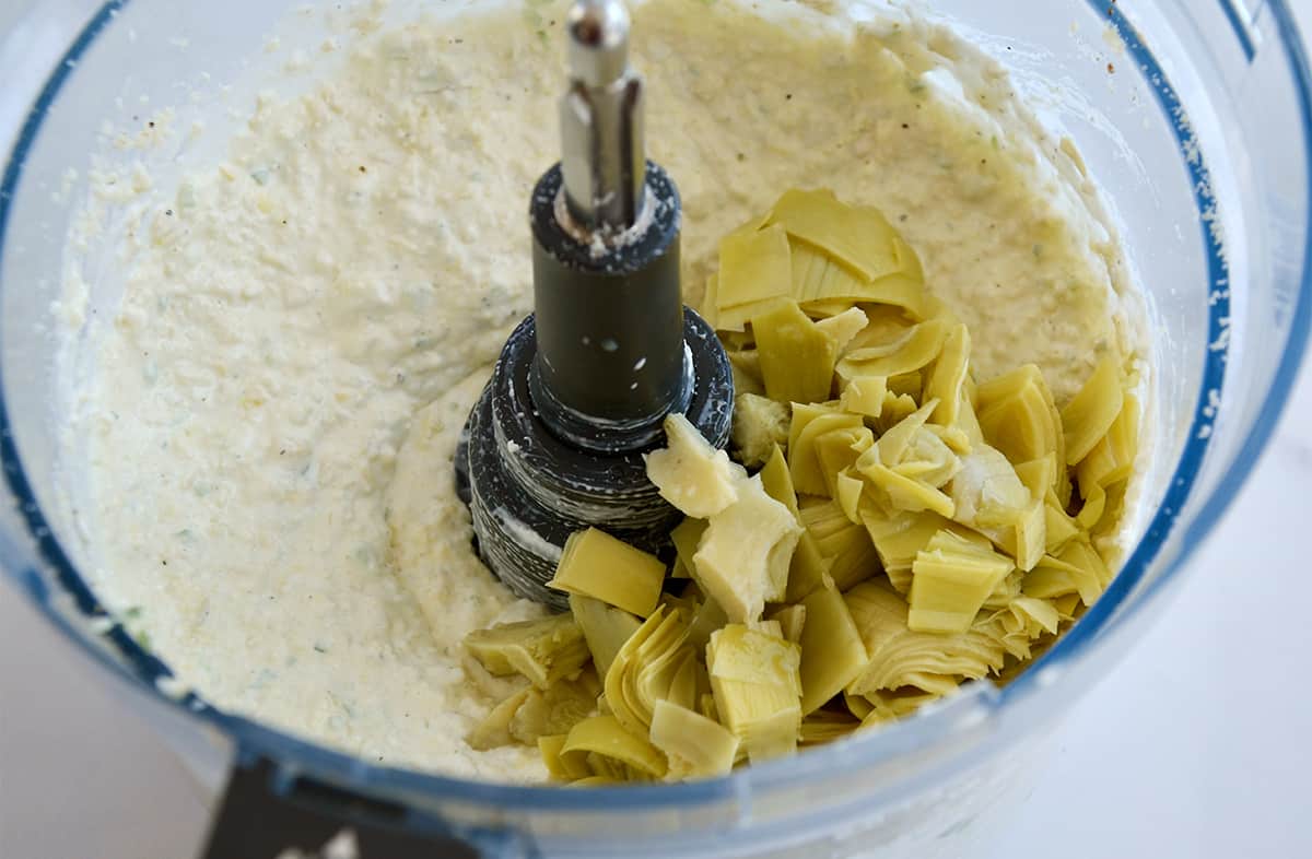 A food processor bowl containing creamy artichoke dip topped with chopped artichoke hearts.