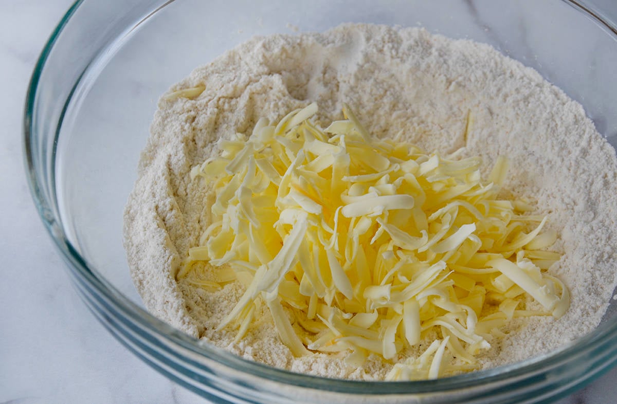 A glass bowl holds grated butter on top of flour.