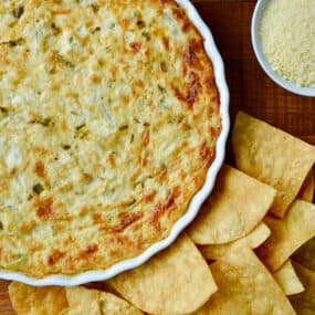 A white baking dish containing warm artichoke dip surrounded by tortilla chips.