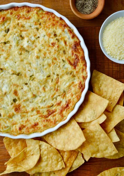 A white baking dish containing warm artichoke dip surrounded by tortilla chips.