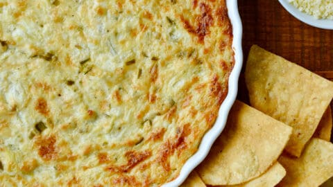 A white baking dish containing warm artichoke dip surrounded by tortilla chips.