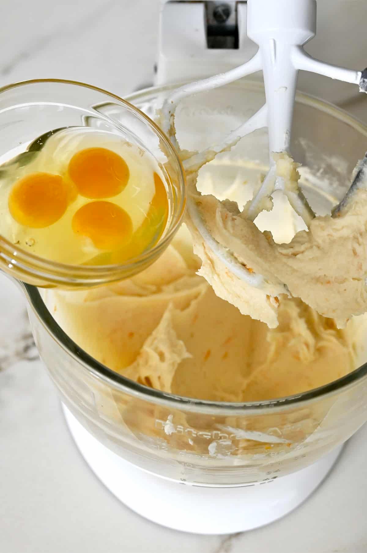 Adding eggs from a small bowl to cream cheese cake batter in a larger bowl.