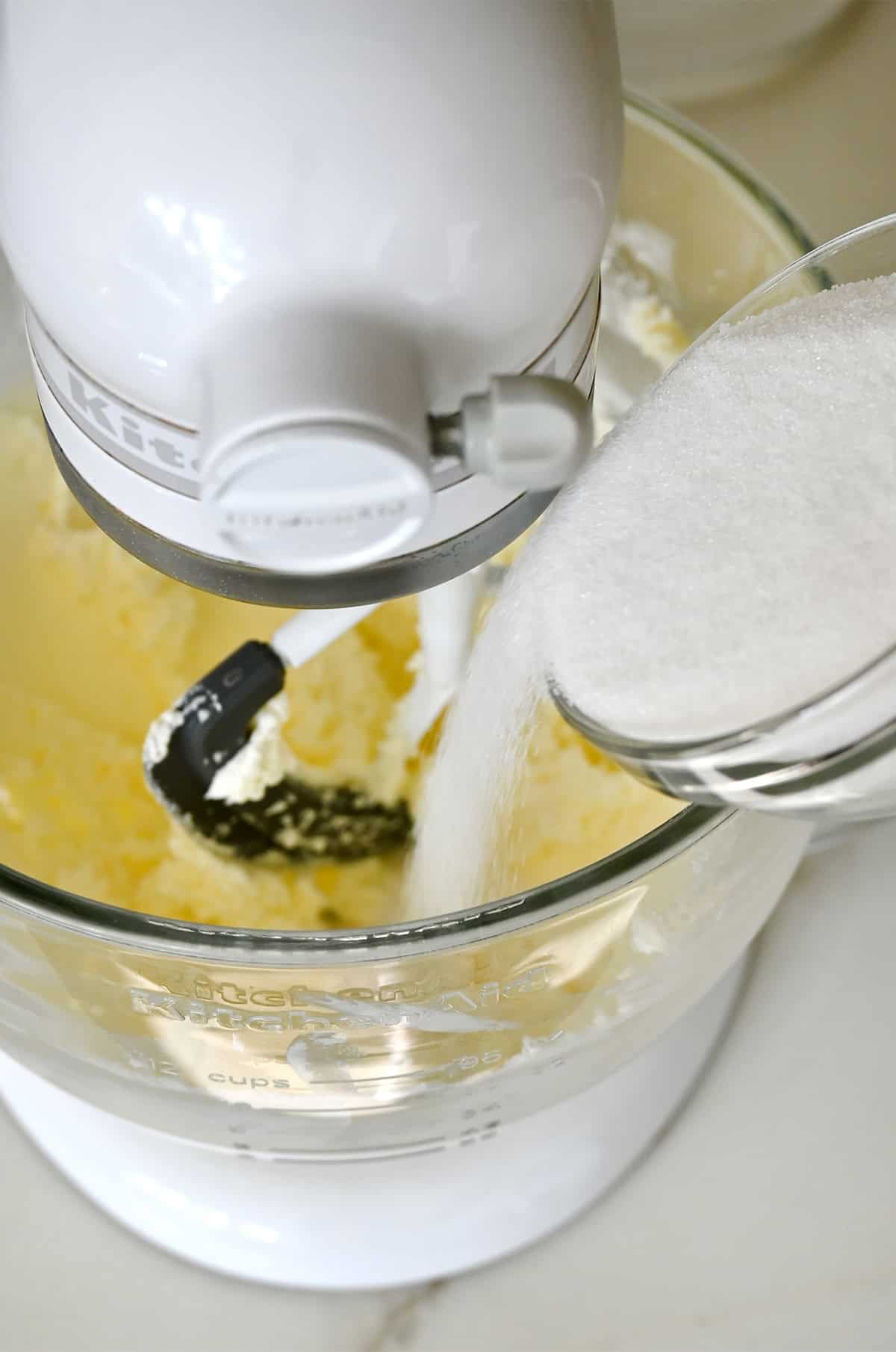 Adding granulated sugar to a stand mixer bowl with creamed butter and cream cheese.