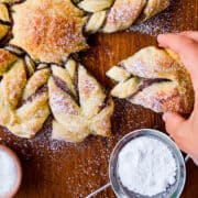 A hand pulling apart a puff pastry snowflake next to. a sifter filled with powdered sugar.