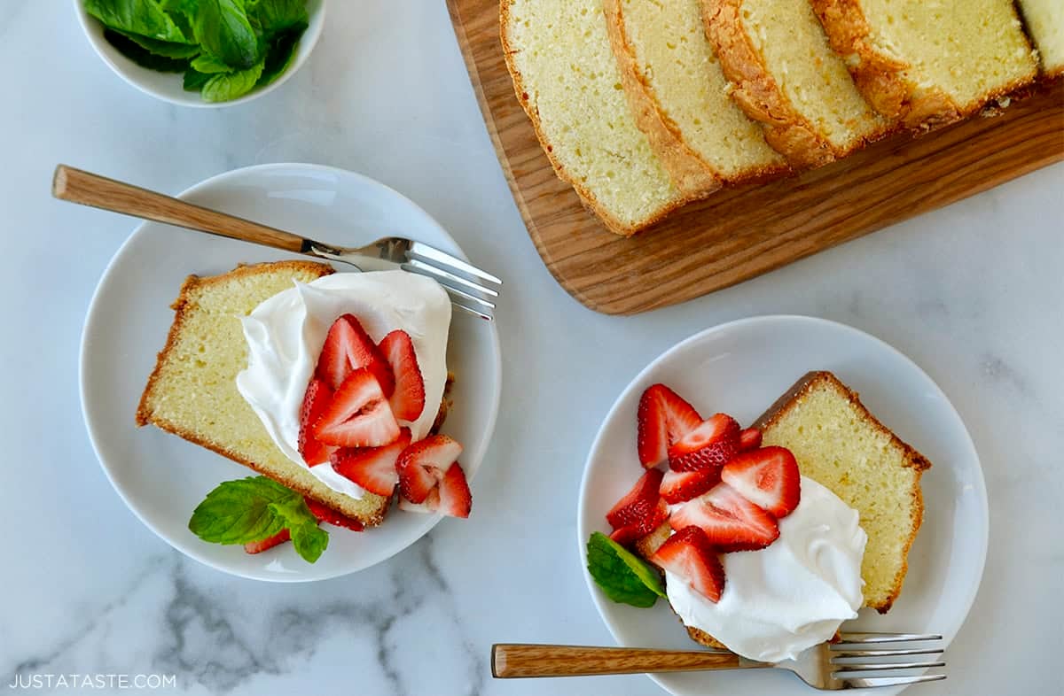 Two dessert plates with a slice of Cream Cheese Pound Cake topped with whipped cream, fresh mint leaves and sliced strawberries next to cutting board with more slices of cake.