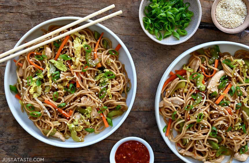 A top-down view of two bowls containing easy chicken chow mein next to small bowls containing sliced scallions, sesame seeds and chili sauce