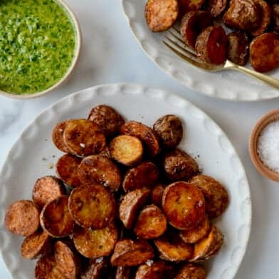 Two plates containing extra crispy baby red potatoes next to a small bowl containing chimichurri sauce.