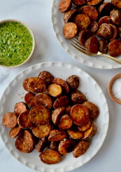 Two plates containing extra crispy baby red potatoes next to a small bowl containing chimichurri sauce.