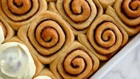 A top-down view of easy Make-Ahead Cinnamon Rolls with Cream Cheese Frosting in a white baking dish next to a bowl of the best cinnamon roll frosting